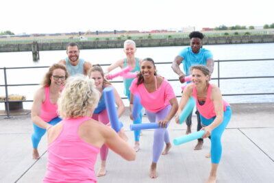 Eine Sportgruppe mit kleinen Poolnudeln als Handgeräten. Die Trainerin steht mit dem Rücken zur Kamera. Im Hintergrund ist die Elbe zu sehen