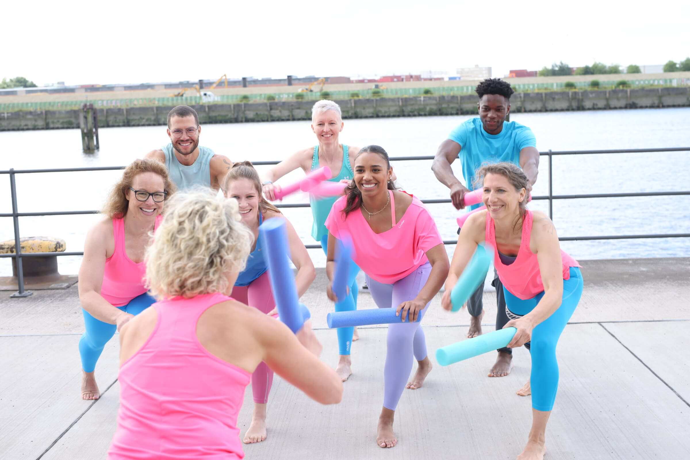 Eine Sportgruppe mit kleinen Poolnudeln als Handgeräten. Die Trainerin steht mit dem Rücken zur Kamera. Im Hintergrund ist die Elbe zu sehen