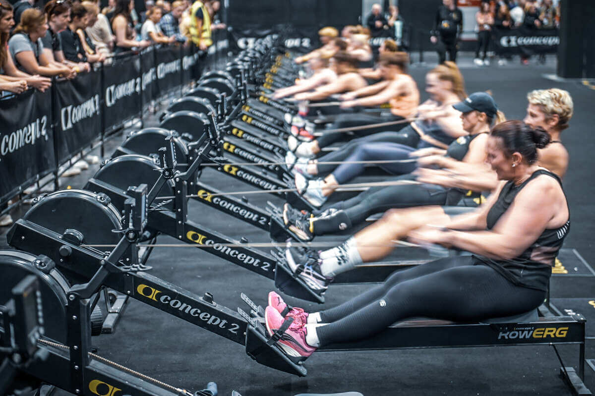 In einer Reihe sitzen Frauen auf einem Fitnessgerät und ziehen an einem Seilzug während eines Hyroxwettkampfes