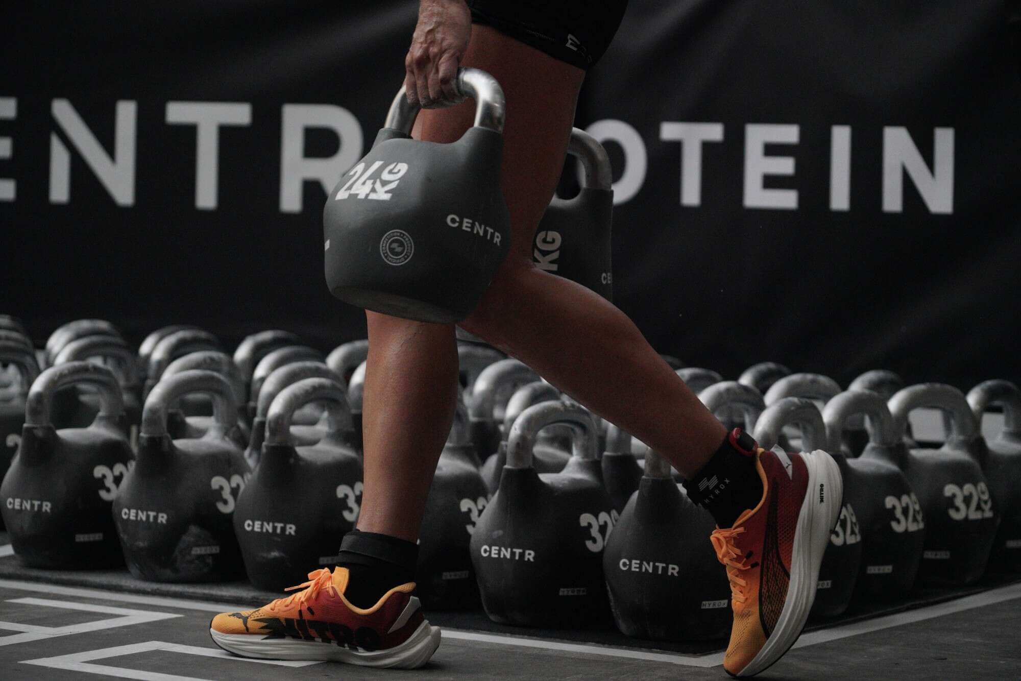 Es ist der untere Körper eine Menschen zu sehen, der in jeder Hand eine 24 KG-Kettlebell trägt. Im Hintergrund stehen sehr viele weitere Kettlebells am Boden.