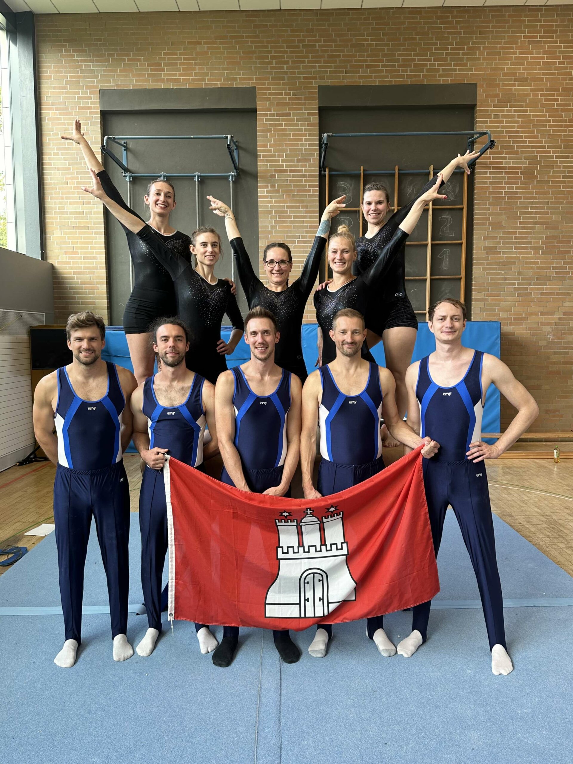 Team Hamburg des Deutschen Senior*innen Pokals Gerätturnen Männer und Frauen im Gruppenbild mit Hamburgflagge. Die Frauen stehen hinter dem Männern erhöht. Alle tragen Turnkleidung