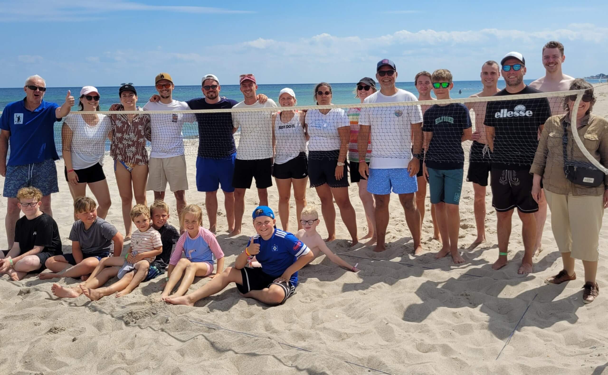 Gruppenfoto aller Teilnehmenden des Beach - Ringtennisturniers am Rosenfelder Strand. Alle haben sich am Netz aufgestellt, einige sitzen davor im Sand. Im Hintergrund ist blauer Himmel und die Ostsee zu sehen.