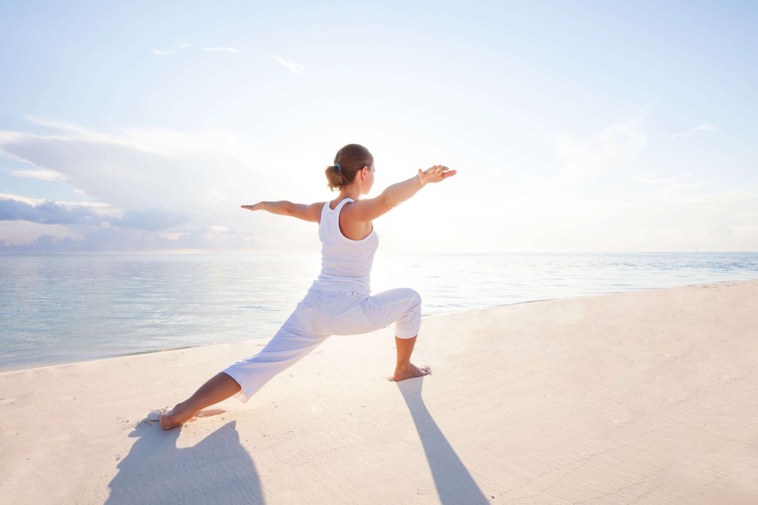 Frau in weißer Sportkleidung macht Yoga am Strand in der Sonne
