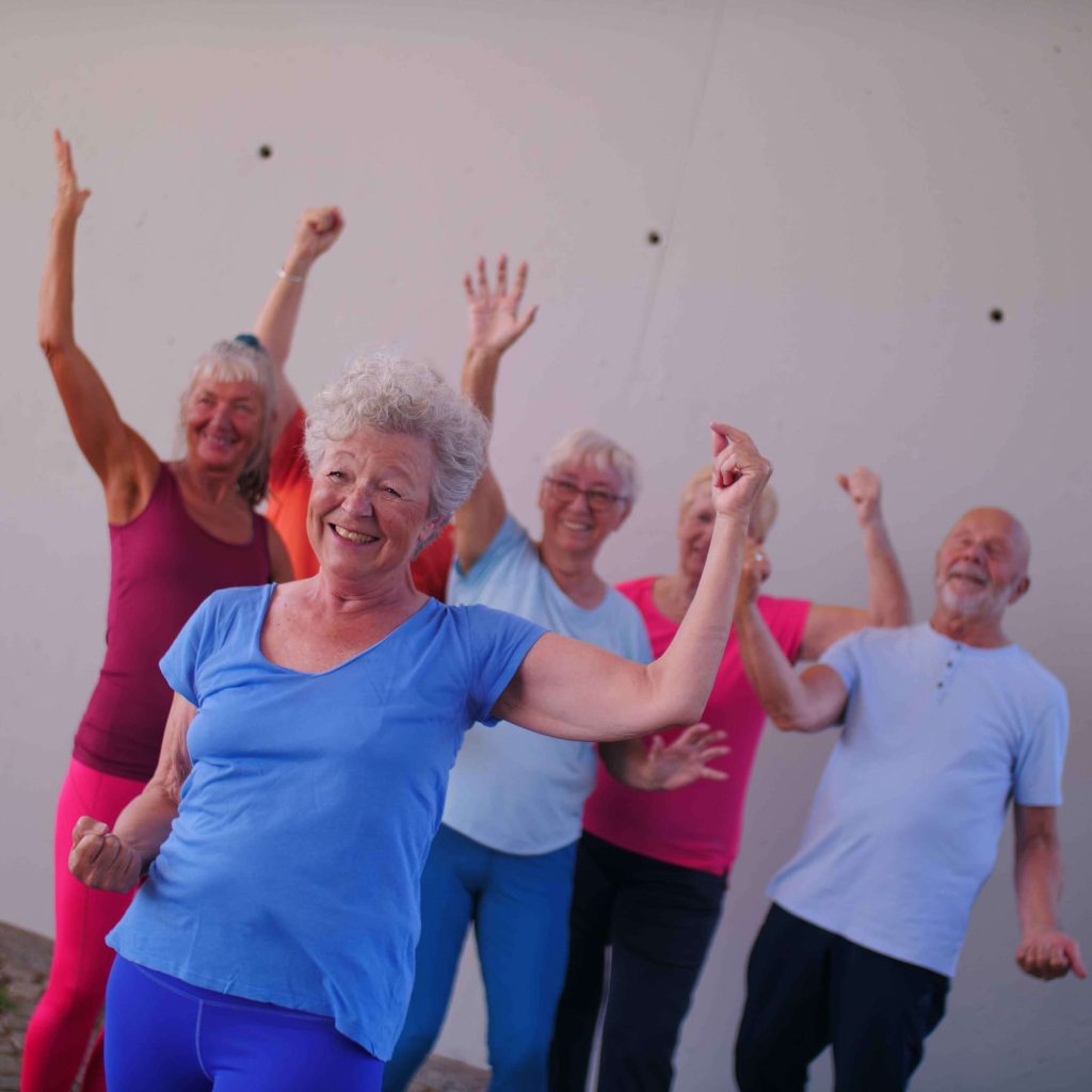 Gruppe älterer Sportler*innen in Fitnesskleidung bewegen sich fröhlich und strecken die Arme in die Luft