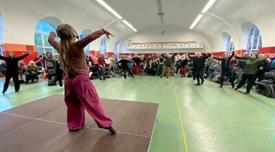 Auf einer Bühne ist eine Frau von hinten zhu sehen, die die Arme für eine Qigong-Bewegung weit ausstreckt. Im weiten Raum vor ihr sind viele Menschen zu sehen, die ihre Bewegung nachmachen.