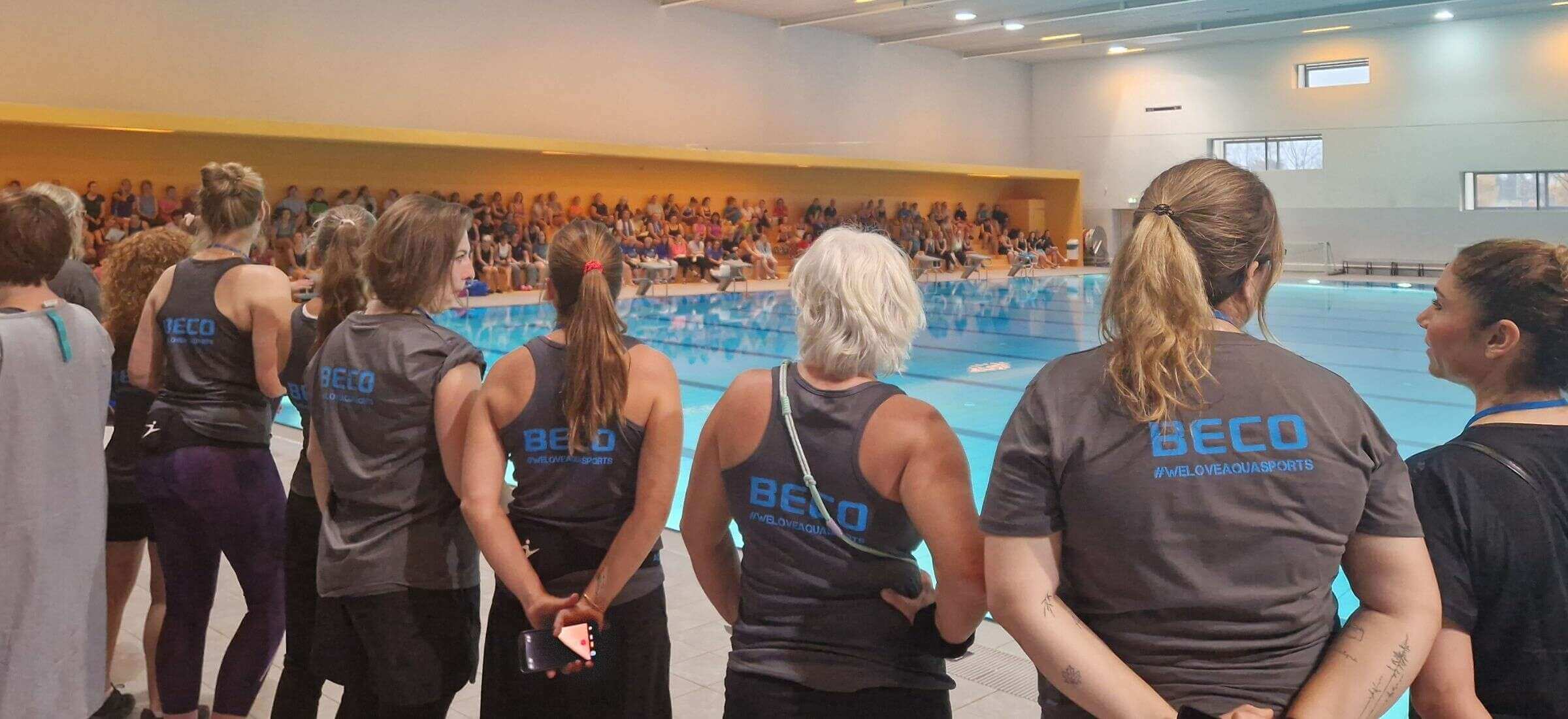 Gruppe erwachsener Frauen stehen mit dem Rücken zur Kamera vor einem Indoor-Schwimmbecken