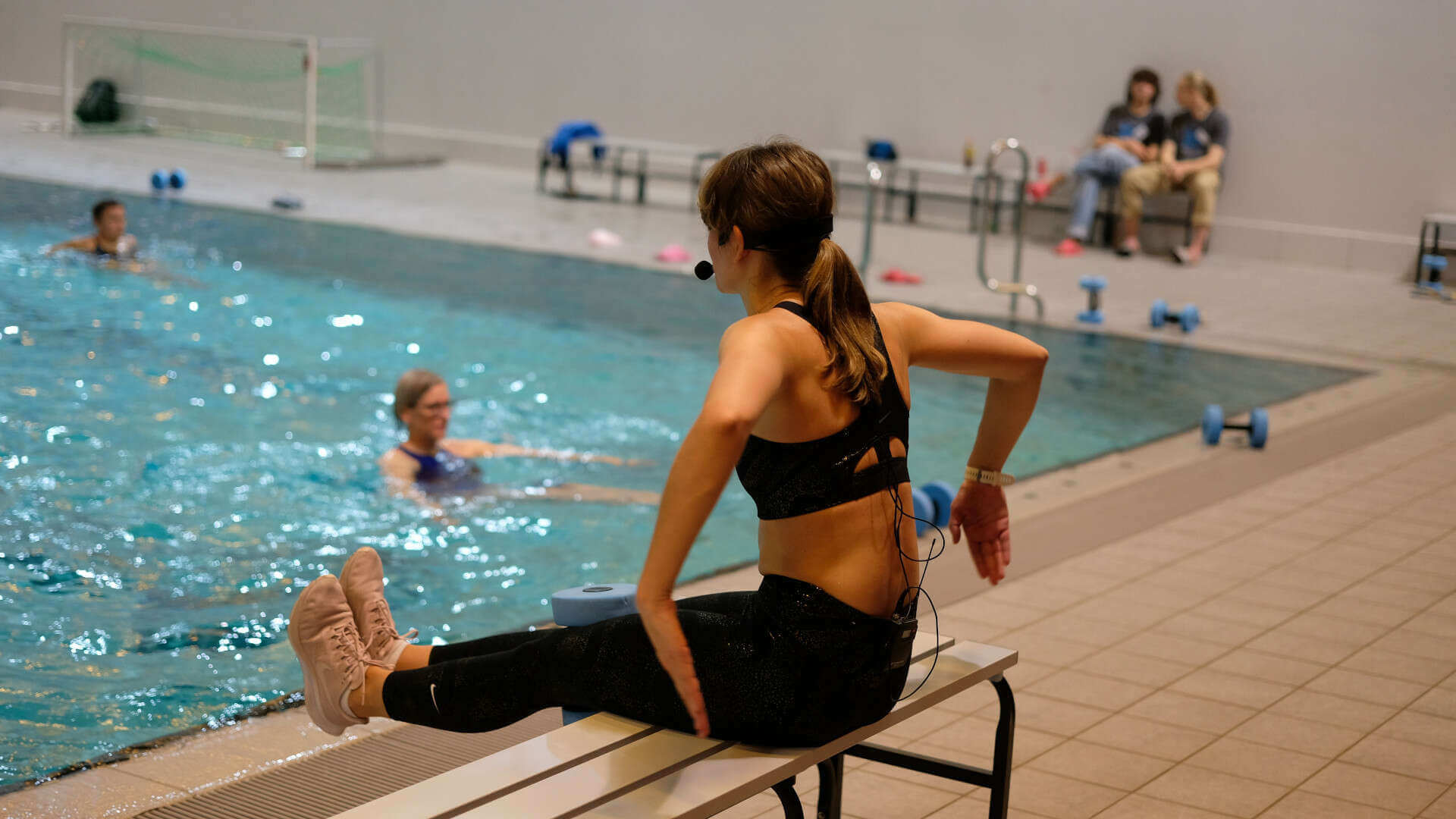 Aqua-Fitness-Trainerin sitzend auf einer Bank mit dem Rücken zum Betrachter, Beine ausgestreckt, Arme seitlich angewinkelt, mit Blick zum Becken, in dem Teilnehmende zu sehen sind.