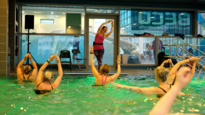 Aqua-Fitness-Trainerin zeigt Übungsgruppe im Wasser eine Übung mit diagonal ausgestrecktem Armen über dem Kopf