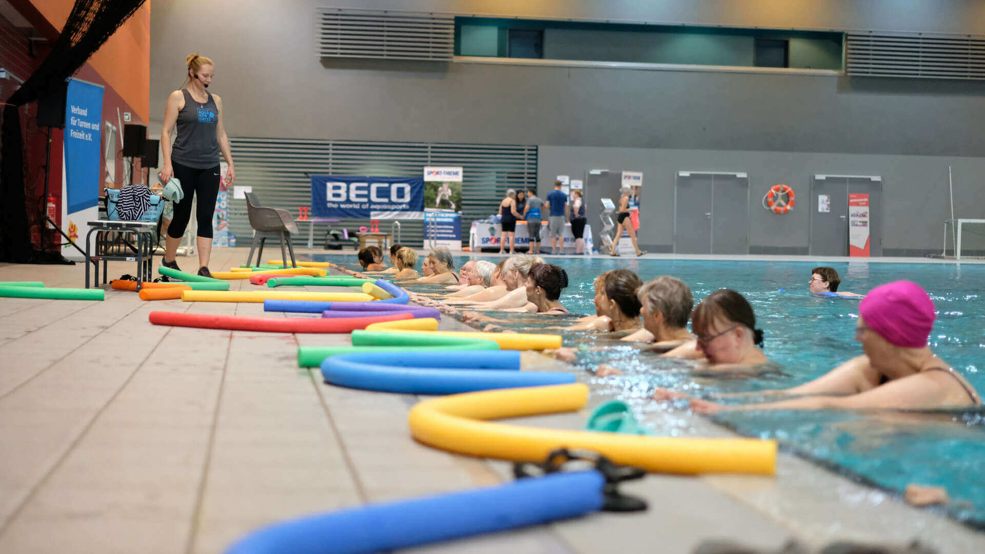 Aqua-Fitnessgruppe am Beckenrand, auf dem Boden liegen bunte Poolnudeln