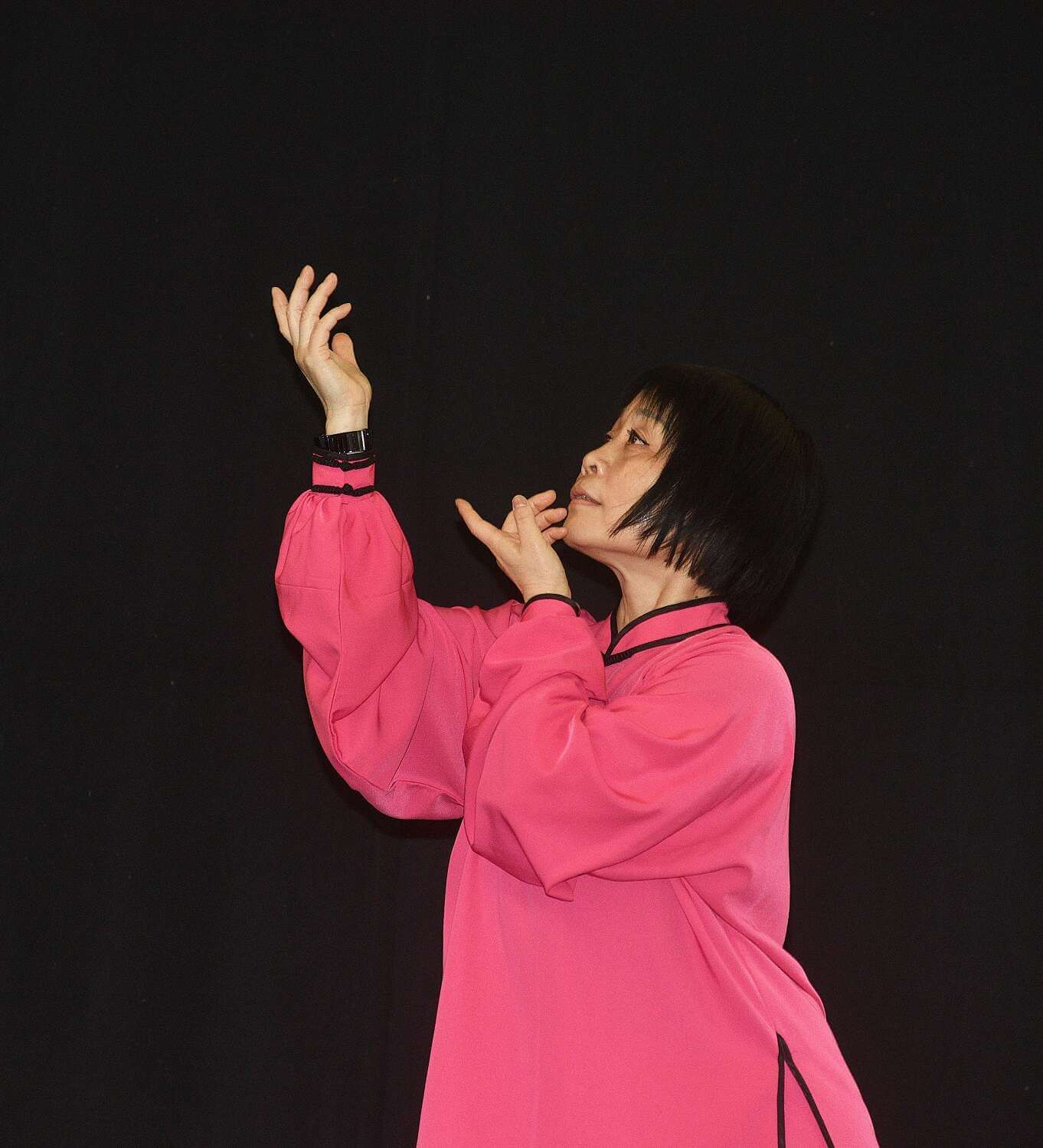 Eine Frau in einem roten Gewand bei einer Bewegung des Qigong, bei der sie beide Hände diagonal mit angewinkelten Armen übereinander nach oben streckt.