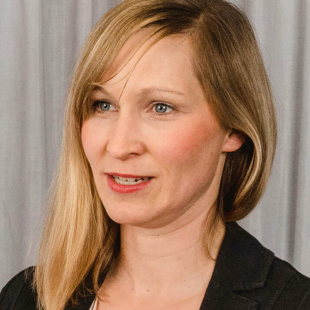 Referentin Anna Breimer trägt schwarzen Blazer, das blonde Haar fällt über die linke Schulter, sie steht vor einem grauen Vorhang