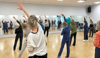In einem Sportraum mit einem Spiegel ist eine Gruppe von Menschen beim Qigong zu sehen. Alle strecken den rechten Arm diagonal nach links oben aus und neigen den Oberkörper leicht nach links