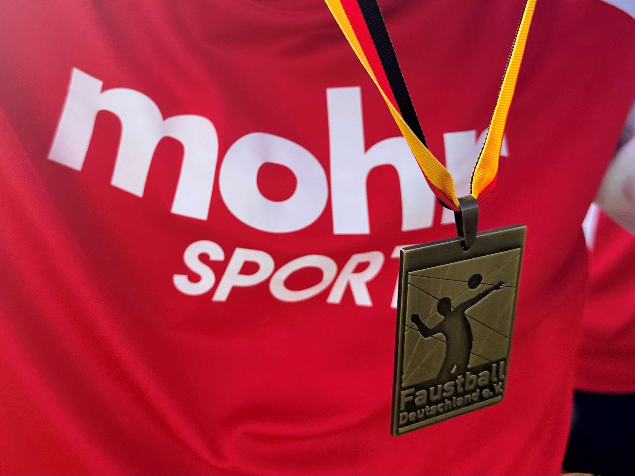 Viereckige Bronzemedaille des Faustball e.V. hängt vor rotem Sporttrikot