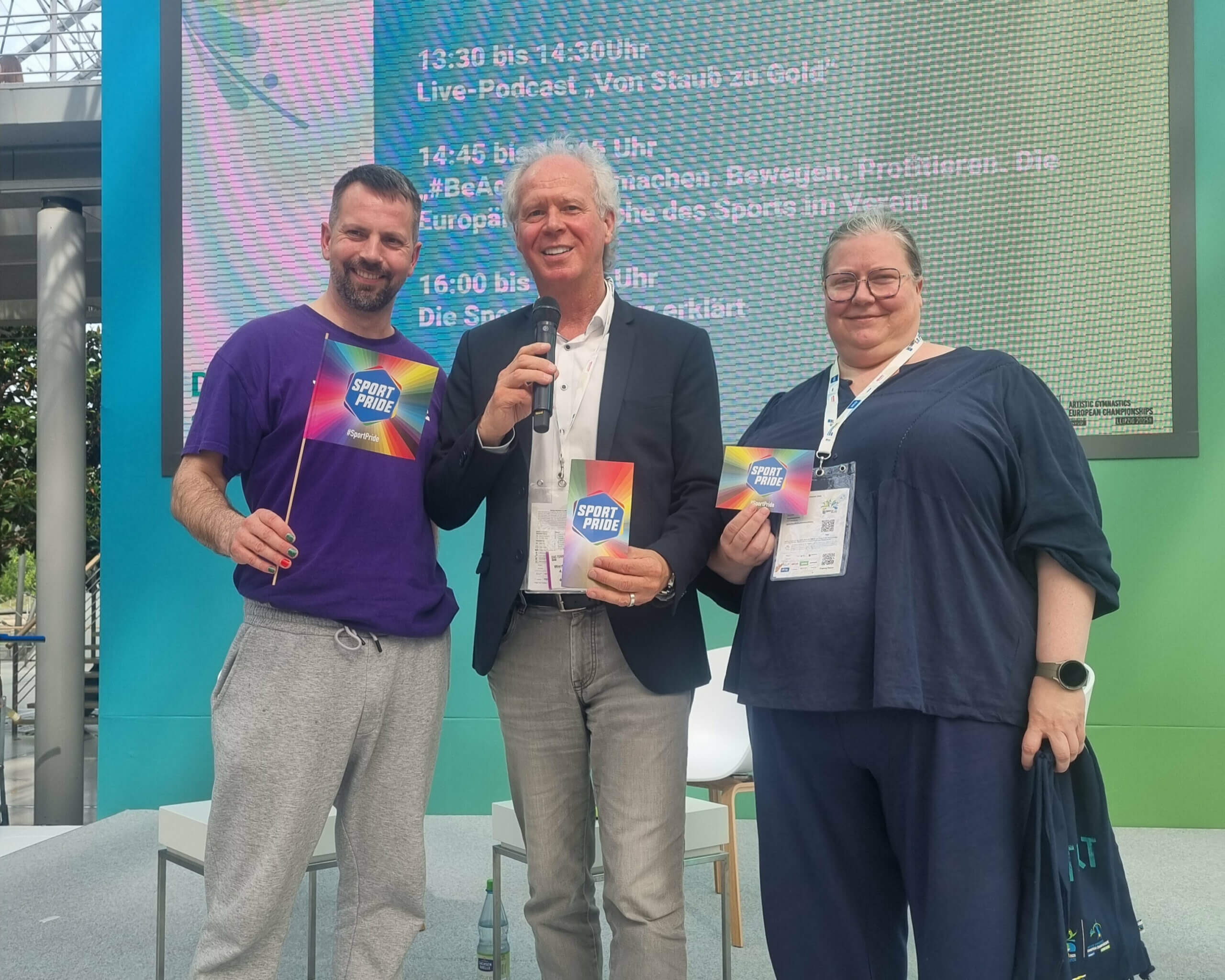 Dörte Kuhn mit zwei Männern auf der Turnfest-Dialogbühne mit SportPride Flagge, Karte und Flyer in den Händen
