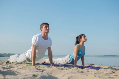 ein Mann und eine Frau machen Yoga am Strand. Sie sind in der Position des "Heraufschauenden Hundes". Im HIntergrund ist das Meer zu sehen.