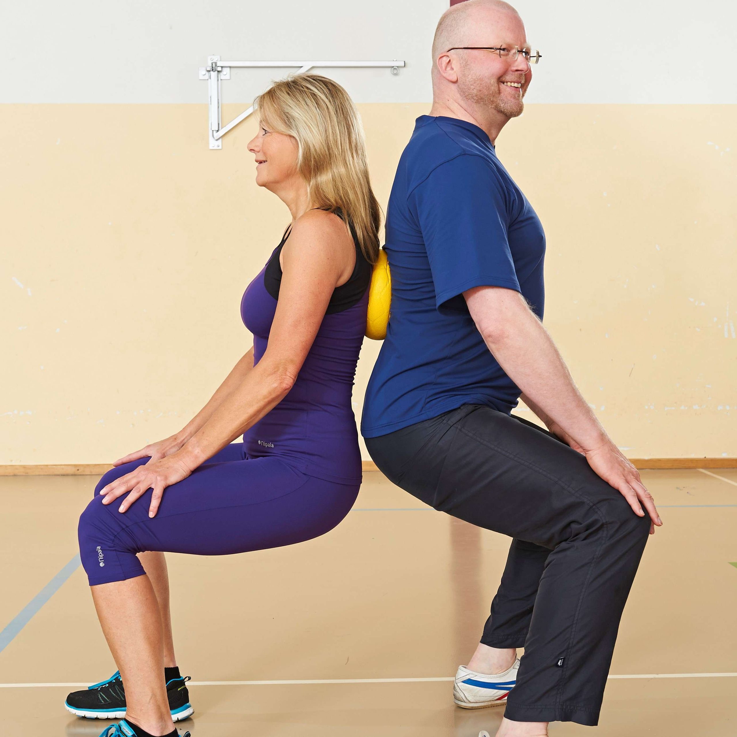 Ein Mann und eine Frau beim Sport stehen in der Kniebeuge Rücken an Rücken dazwischen ist ein gelber Ball eingeklemmt.