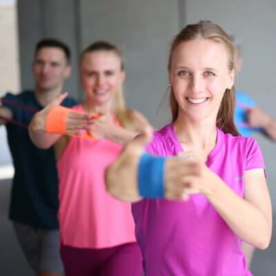 Frau in pink farbenen T-Shirt streckt die Hand mit einem Thera-Band nach vorne. Im Hintergrund stehen drei weitere Personen in Fitnesskleidung, die es nachmachen.
