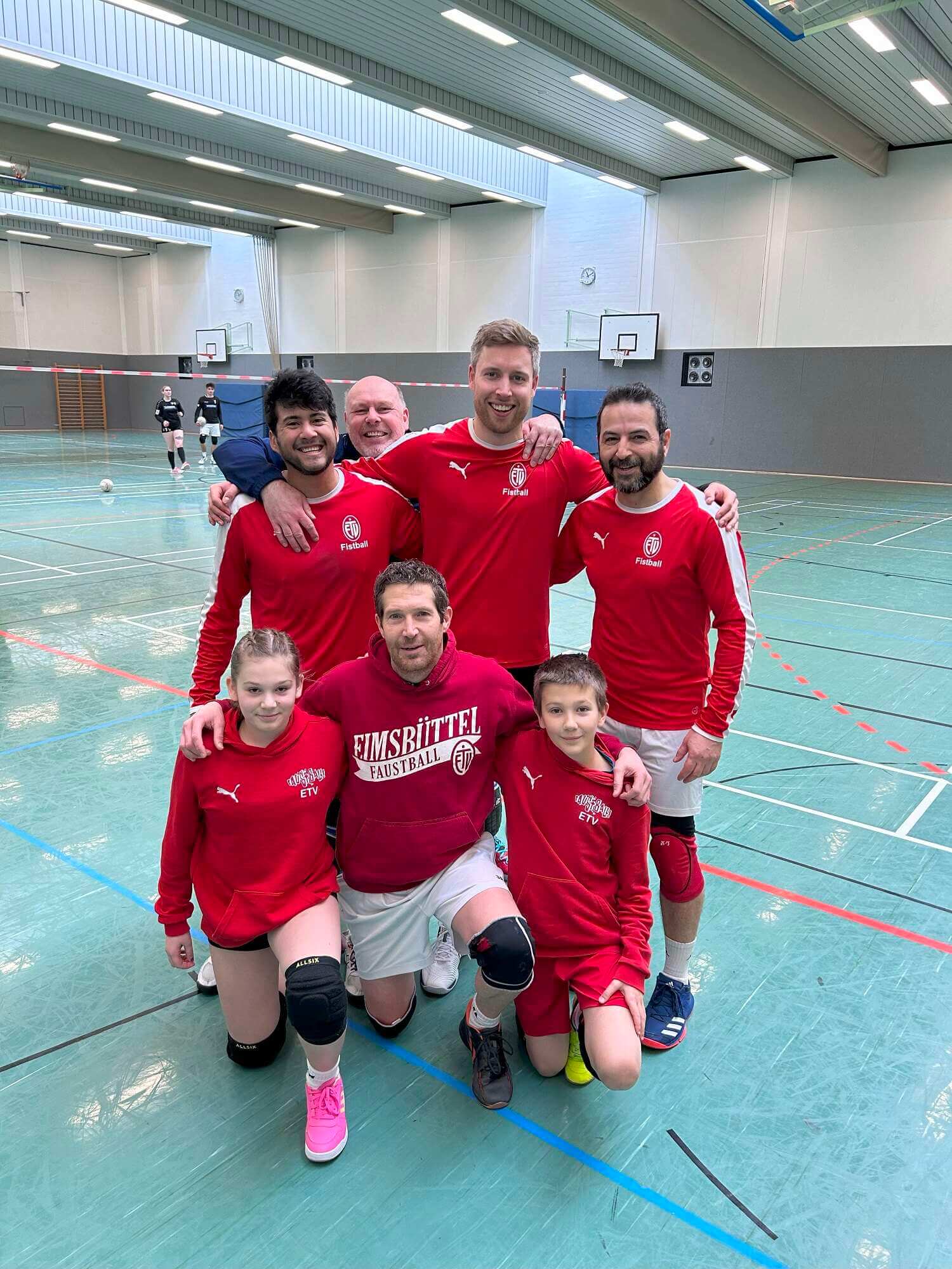 Ein Mannschaftsfoto der Faustballmannschaft des GFSV Hamburg, bestehend aus 5 Männern und zwei jugendlichem, ein Mädchen und ein Junge. Alle tragen ein rot-weißes Trikot, einer ein schwarzes Sportoutfit.