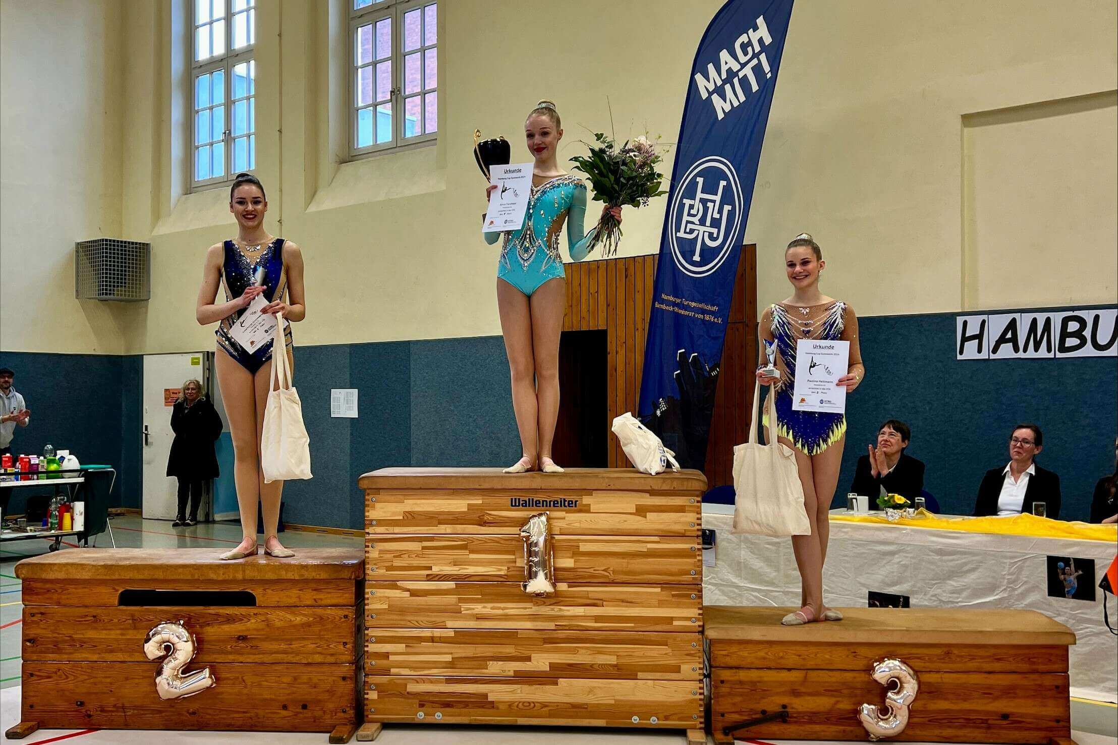 Drei Gymnastinnen stehen auf den Plätzen 1-3 auf dem Siegerpodest, alle halte eine Urkunde und einen Podkal in der Hand, die Siegerin hält zudem einen Blumenstrauß