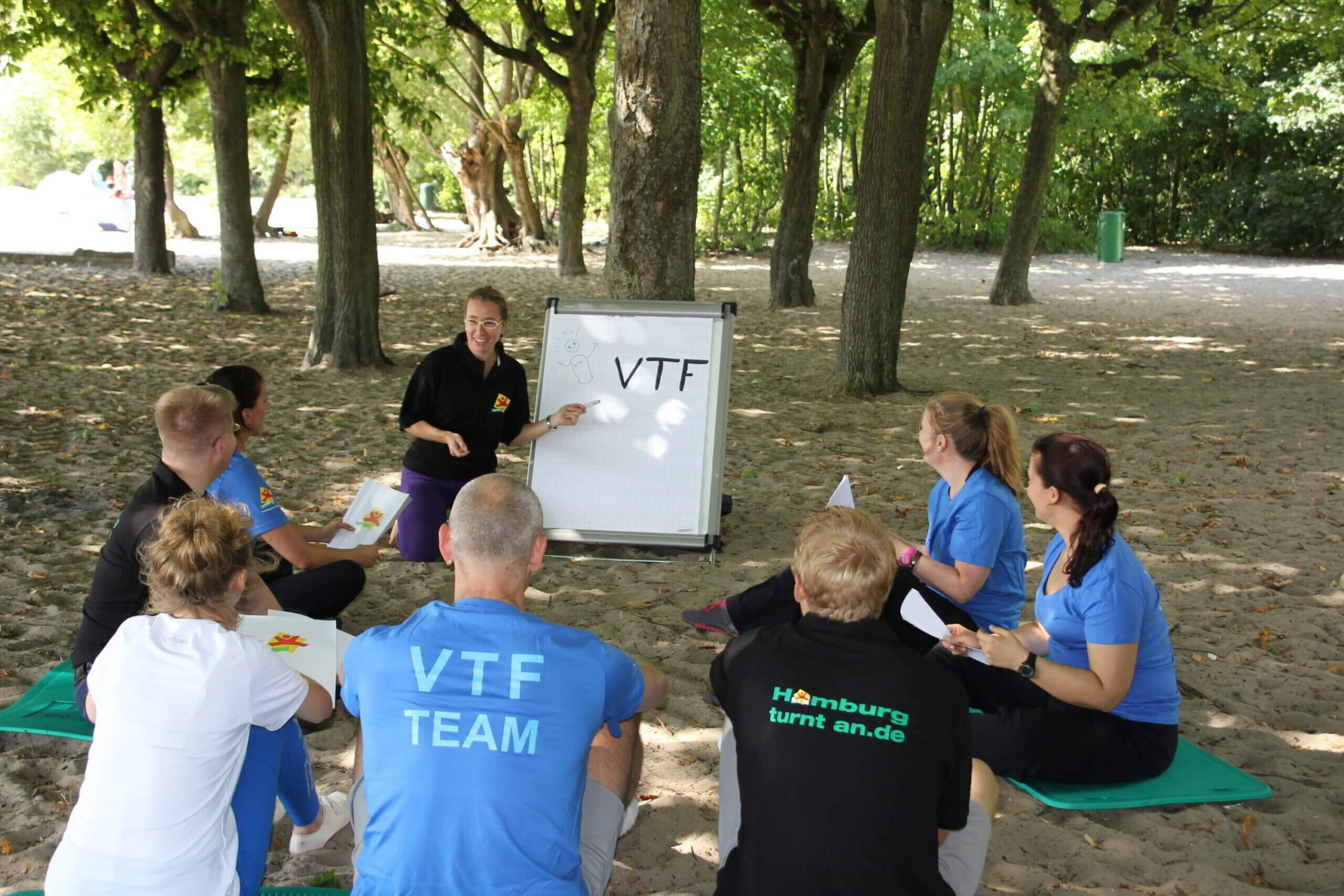 ein bundesfreiwilligen Dienst im Kreise des VTF-Team bringt Spaß. Man sieht eine Gruppe von Personen aus dem VTF-Team, deren T-Shirts sie tragen, sitzen in einem Kreis auf Gymnastikmatten im Sand. Eine Frau zeigt auf ein Flipchart, auf dem VTF steht.