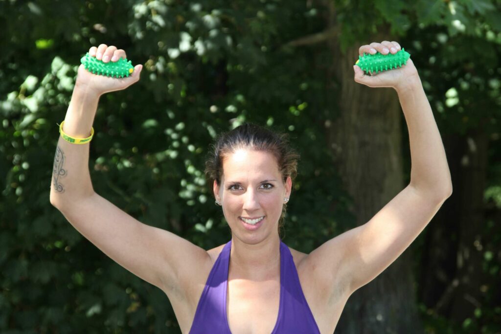 Eine Frau in Sportkleidung, deren Oberköper. Kopf und Arme zu sehen sind, hält die Arme über dem Kopf, in beiden Händen hält sie jeweils Brazils (ein Handgroßes grünes Fitnessgerät)