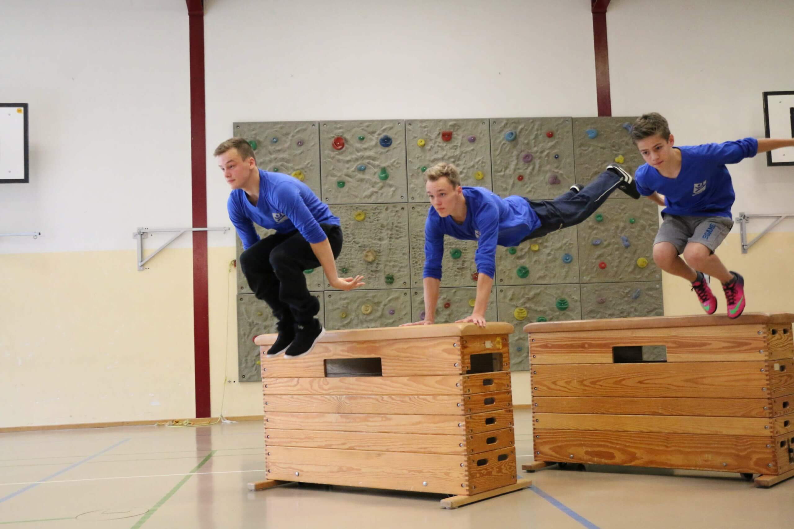 Drei Jungen in blauen T-Shirts springen mit einer Hocke über zwei Turnkasten