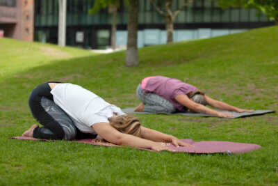 Zwei Frauen mit Ihren Yogamatten auf einer Wiese mit der Übung Ardha-Kurmasana