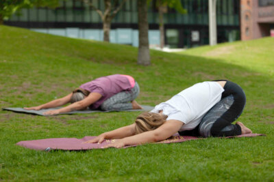 Zwei Frauen mit Ihren Yogamatten auf einer Wiese mit der Übung Ardha-Kurmasana