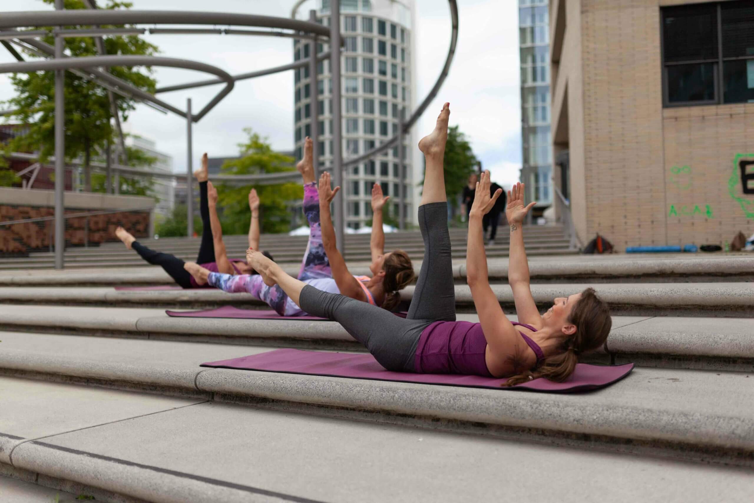 Drei Frauen liegen versetzt auf drei Steinstufen hintereinander auf ihren Yogamatten. Sie spreizen ein Bein in die Luft, und beide Arme sind ebenfalls nach oben gestreckt. Im Hintergrund ist ein Hochhaus zu sehen.