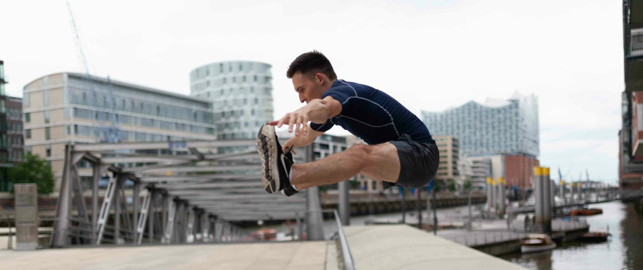 Ein Sportler springt im freien Grätschsprung über ein Geländer. Im Hintergrund ist die Hafencity Hamburgs zu sehen.