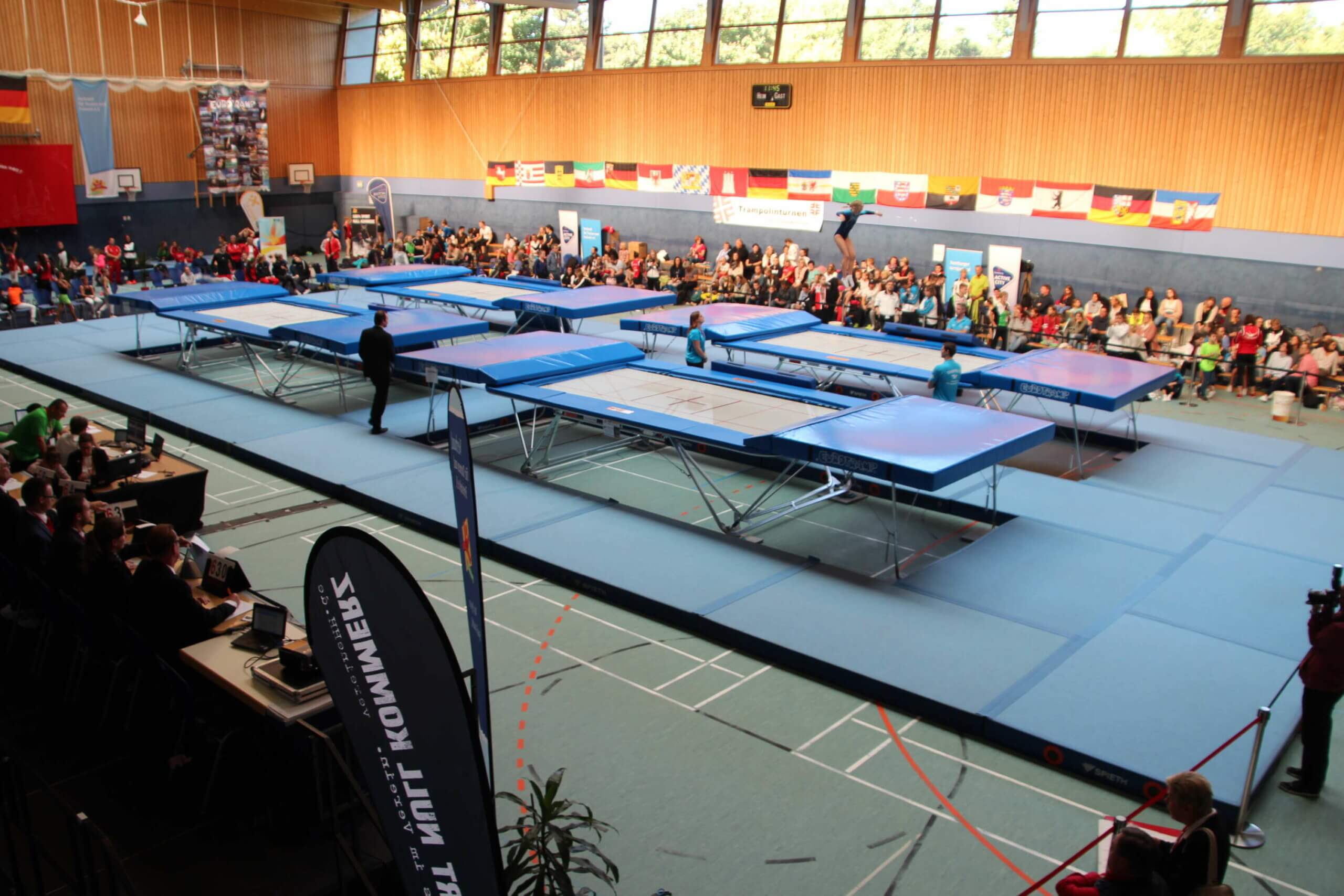 In einer Sporthalle stehen inmitten einem riesigen Mattenkreis 4 große Trampoline. Runterum an den Hallenwänden sind Bundesländerfahnen aufgehängt