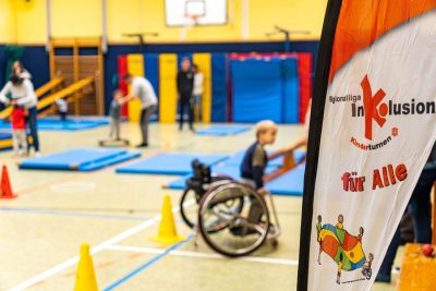 Im Vordergrund ist eine Fahne zu sehen, auf der Inklusion für alle steht. Im Hintergrund sieht man eine Turnhalle mit verschiedenen Übungsstationen. Unter den Kindern sieht man auch einen Jungen im Rollstuhl.