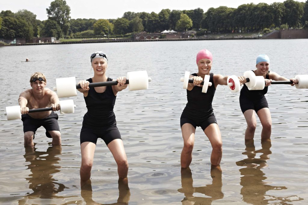 Vier Aqua-Fitness-Sportler*innen mit einer Wasserhantel stehen in einer Kniebeuge im wadentiefen Wasser eines Sees. In der Hand haben sie in Vorhalte eine Schwimmhantel. Die drei Frauen tragen eine Badekappe und einen traditionellen Ganzkörperschwimmanzug. Der Mann trögt eine schwarze, knielange Badehose