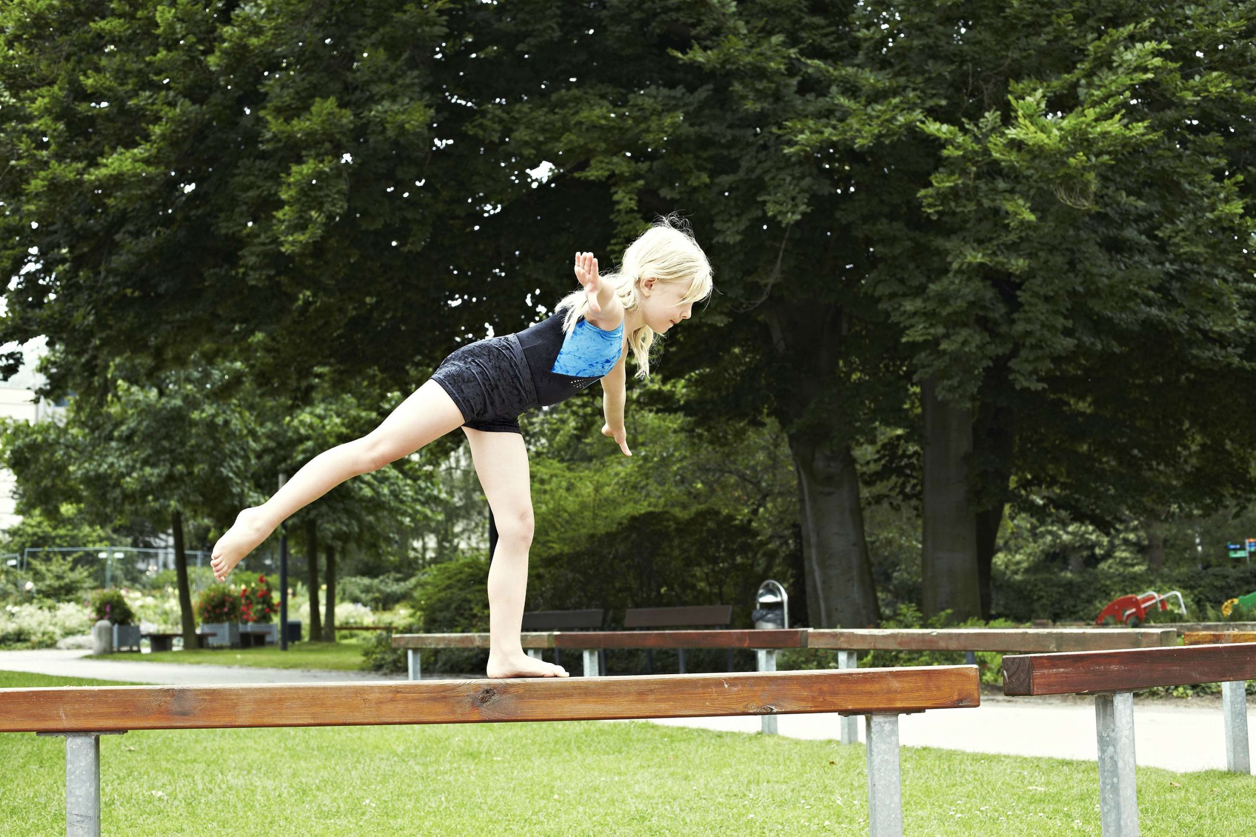 Ein Mädchen steht auf einem hölzernen Schwebebalken im Turnanzug balanciert auf einem Bein, das andere Bein ist nach hinten ausgestreckt