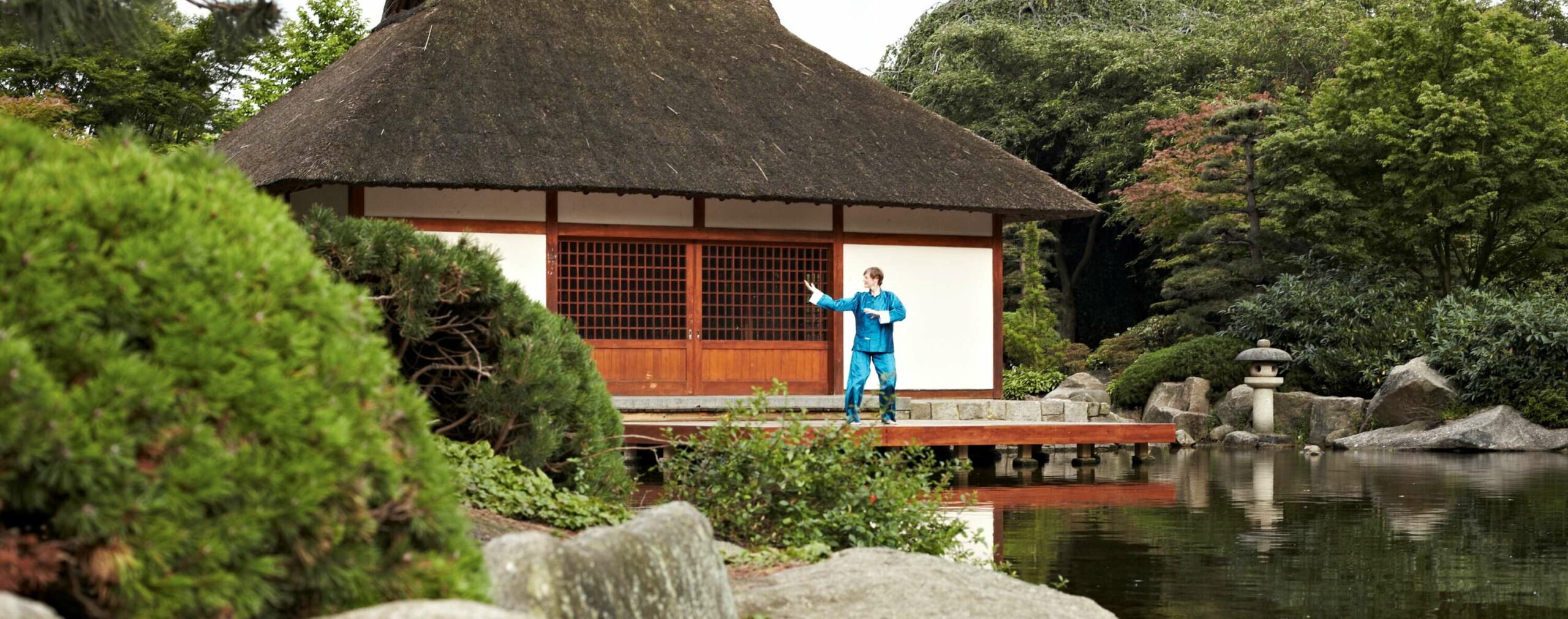 Person im blauen Seidenanzug beim Qigong vor einem Pavillon am Wasser