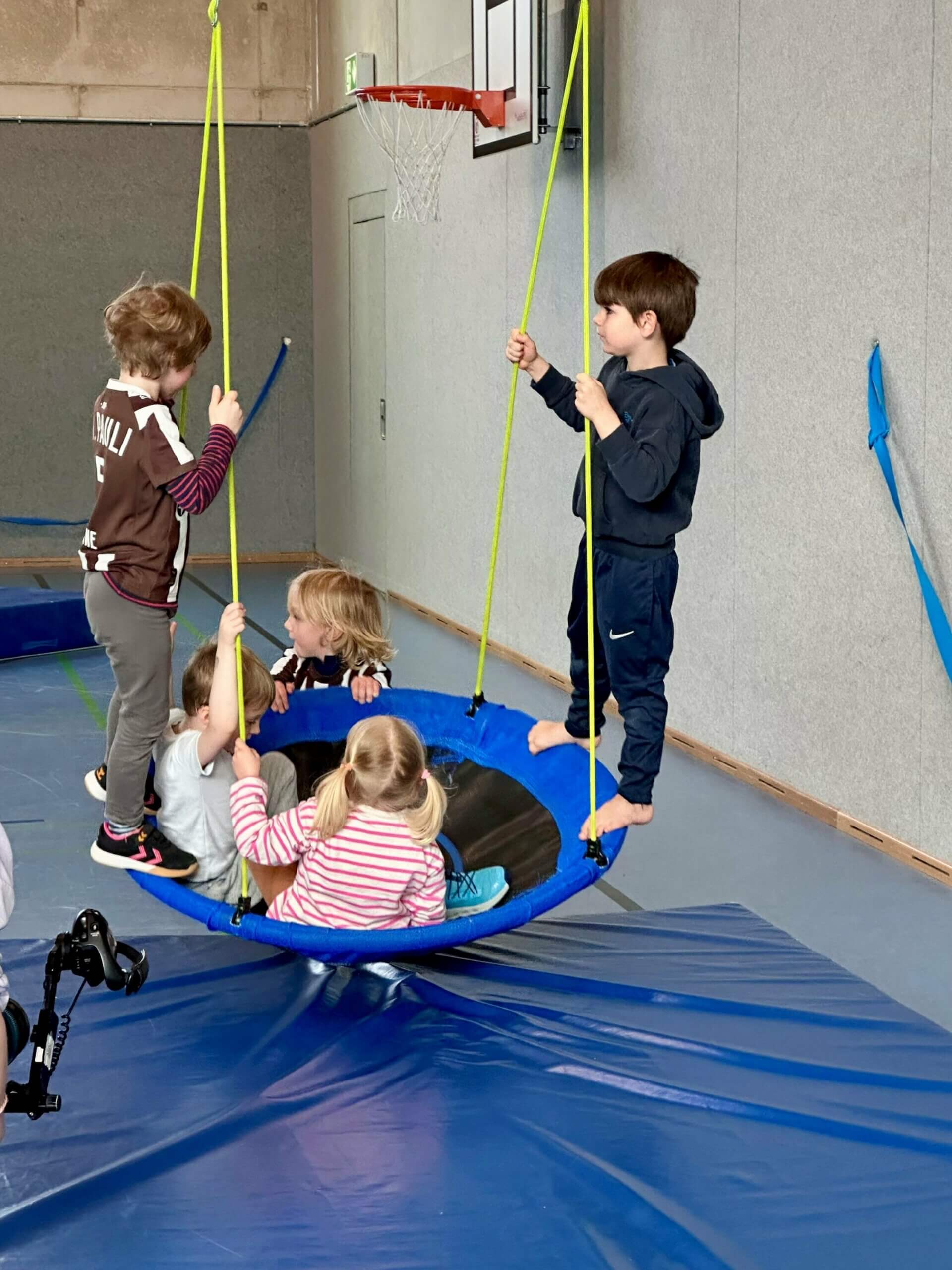 Eine Gruppe kleinerer Kinder schwingt auf einer großen Korbschaukel in einer Turnhalle. Zwei Stehen am Rand und drei Kinder sitzen darin.