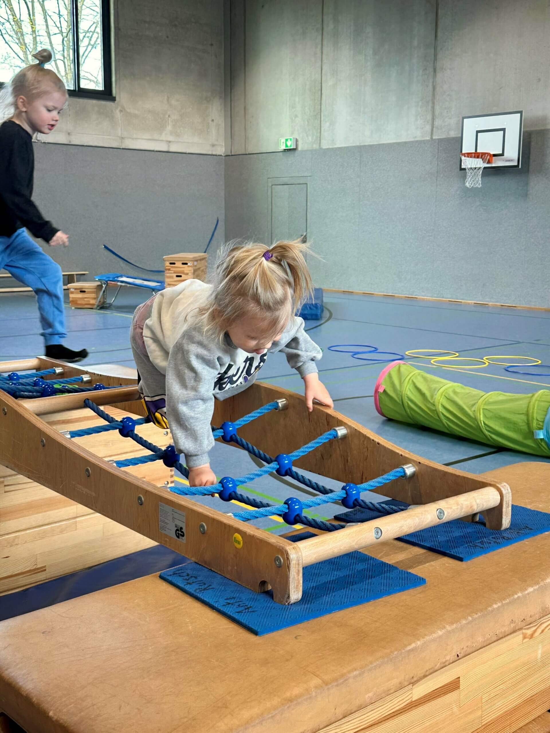 Ein Kind beim Kinderturnen klettert über eine horizontal liegende Leiter aus Tauen.