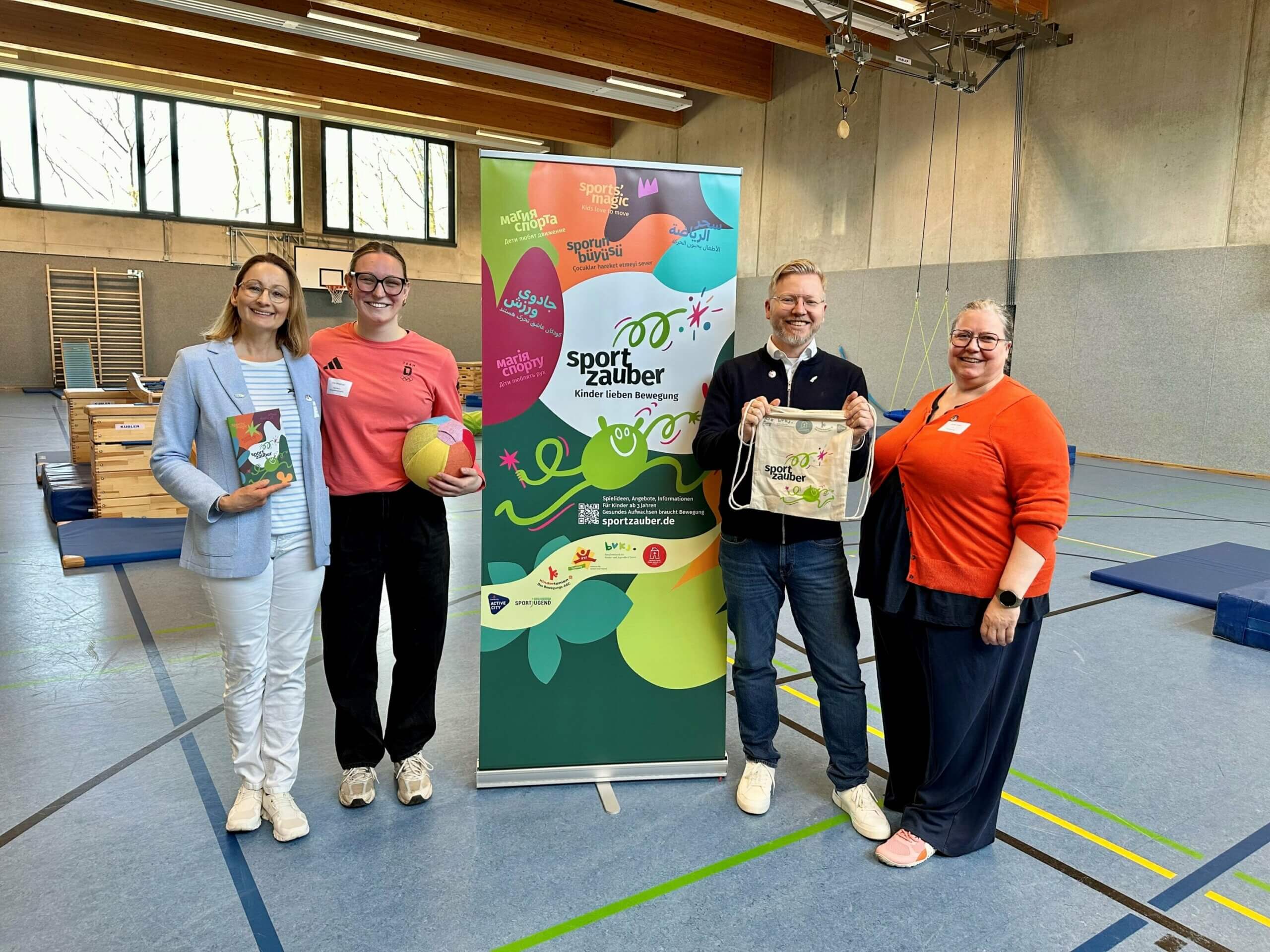 Ein bunter Aufsteller mit dem Logo Sportzauber steht in der Mitte von 4 Personen, sie halten eine Broschüre, einen Turnbeutel und einen bunten Ball in Höhe, im Hintergrund ist eine Turnhalle mit Geräten zu sehen.