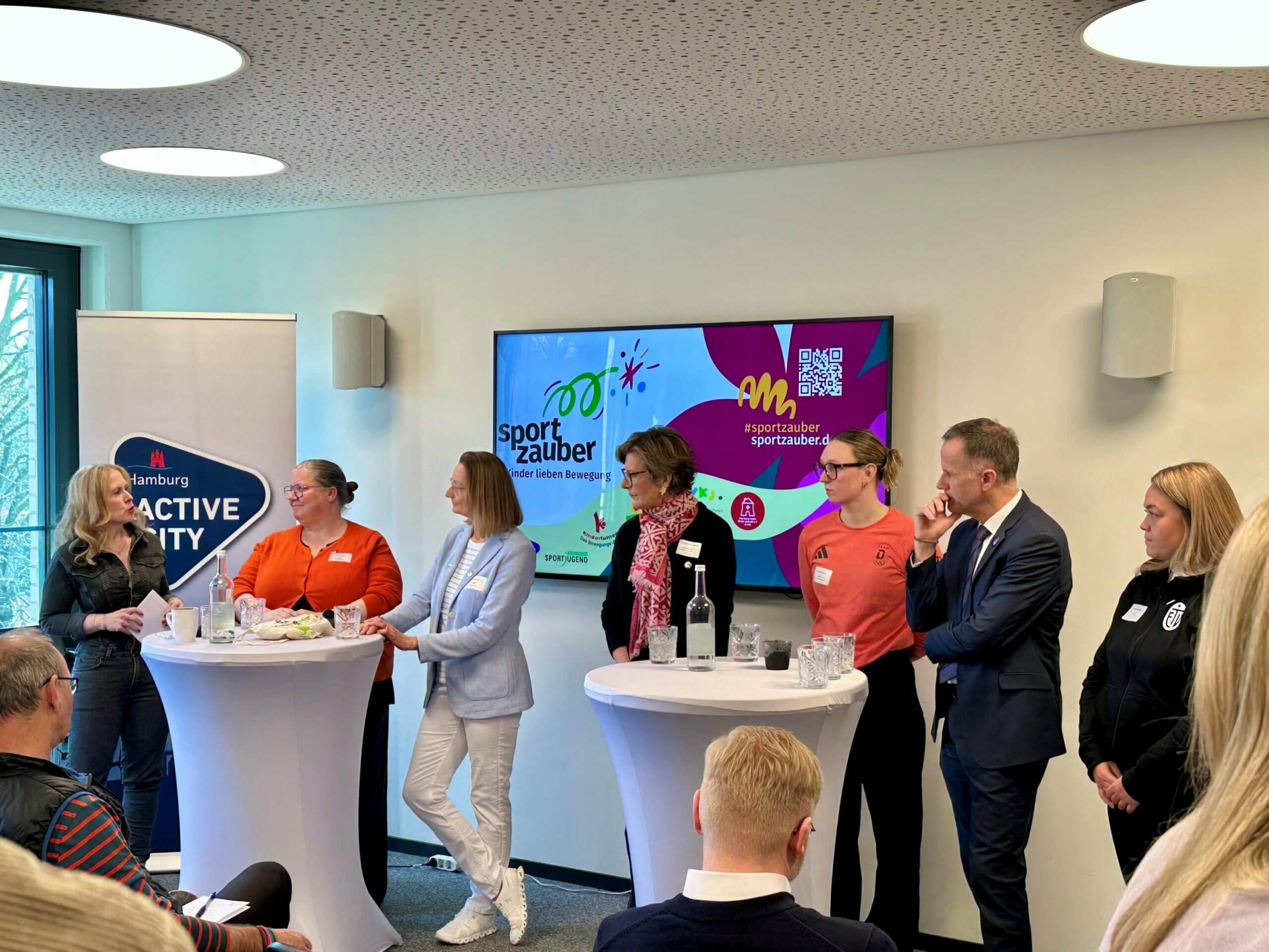 Pressegespräch Sportzauber. Es stehen 7 Redner*innen hinter zwei Stehtischen. Im Hintergrund ist das bunte Bild und der Schriftzug von Sportzauber auf einem großen Bildschirm zu sehen