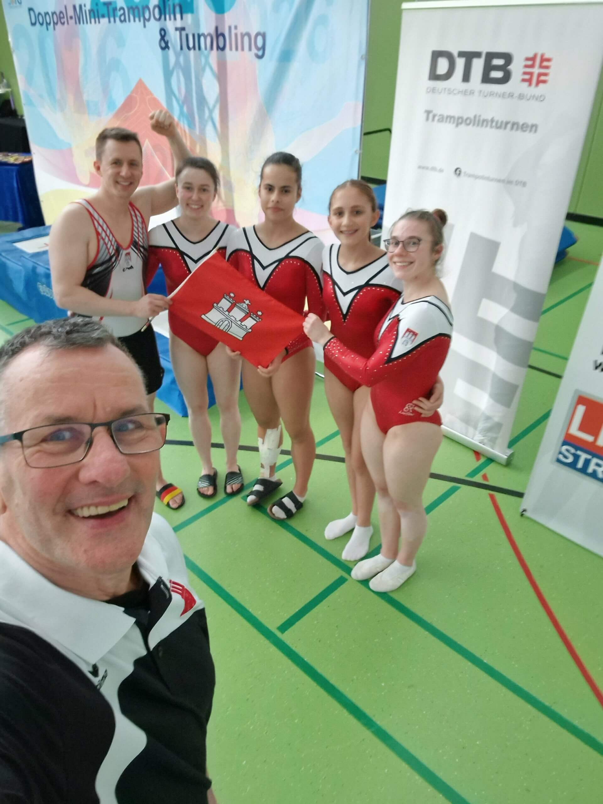 Trampoliner*innen in roten Anzügen mit ihrem Trainer in der linken Ecke