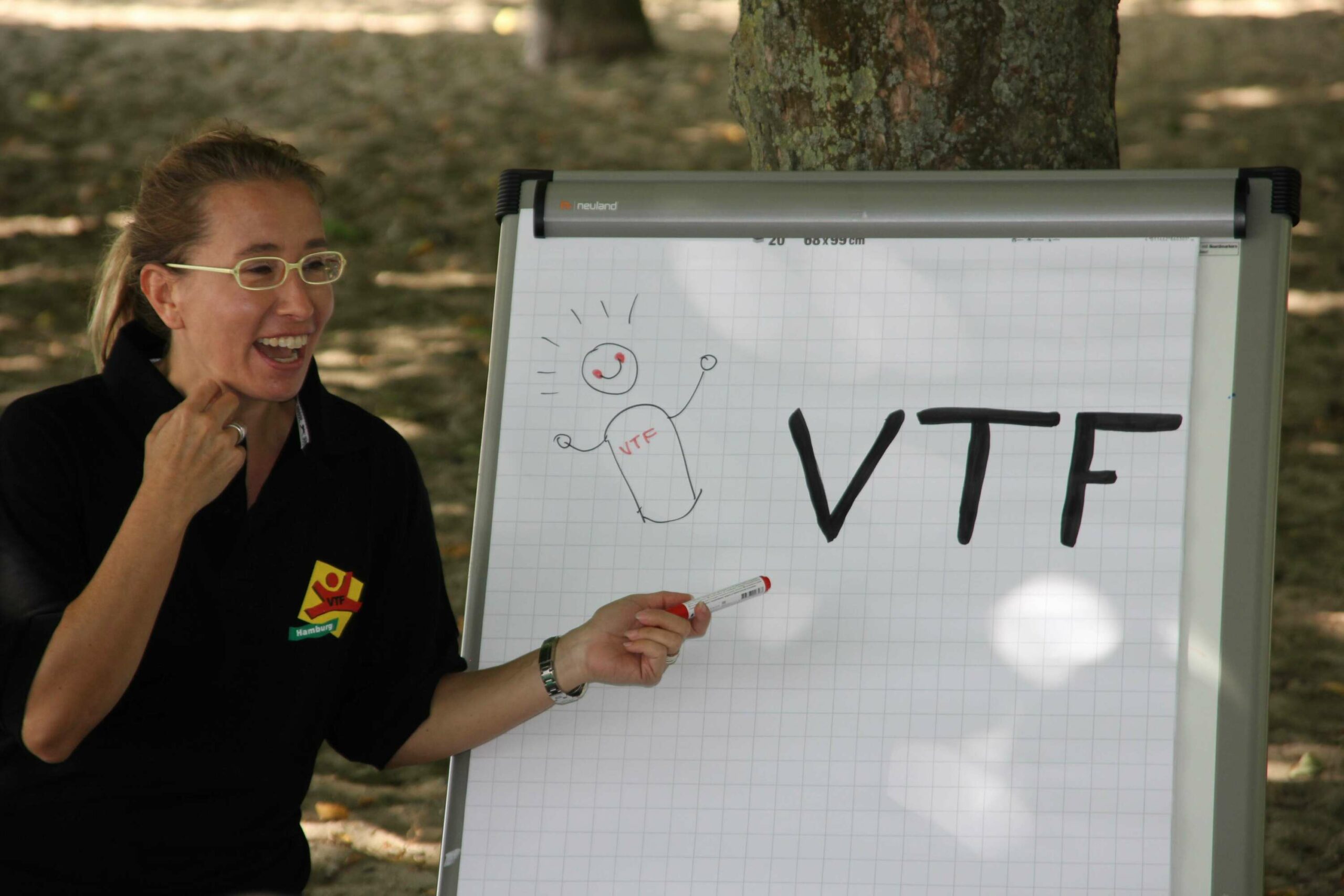 Frau in einem schwarzen T-Shirt, zeigt mit einem Stift auf den Schriftzug VTF auf einem Flipchart