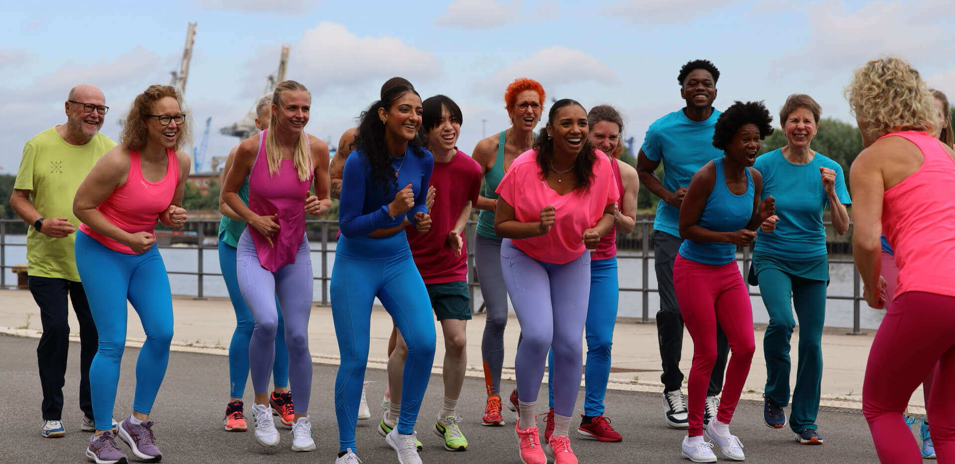 Eine Gruppe von Fitnesssportler*innen in einer läuferischen Haltung auf der Stelle mit euphorischen, begeisterten Gesichtern. rechts vorne im Bild ist die Trainerin von hinten zu erkennen, sie macht die Bewegung vor.