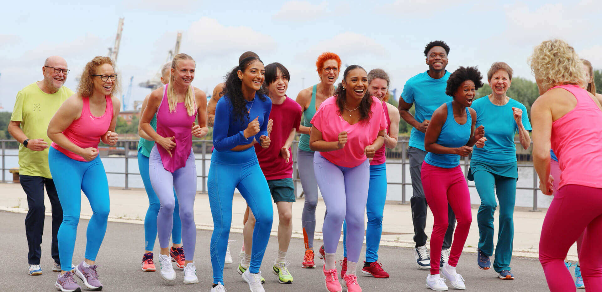 Eine Gruppe von Fitnesssportler*innen in einer läuferischen Haltung auf der Stelle mit euphorischen, begeisterten Gesichtern. rechts vorne im Bild ist die Trainerin von hinten zu erkennen, sie macht die Bewegung vor.