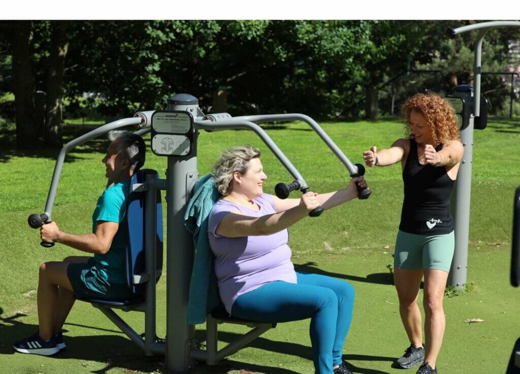 Frau auf einem Outdoor Sportgerät, lächelt. Zweite Frau zeigt ihr korrekte Nutzung des Geräts