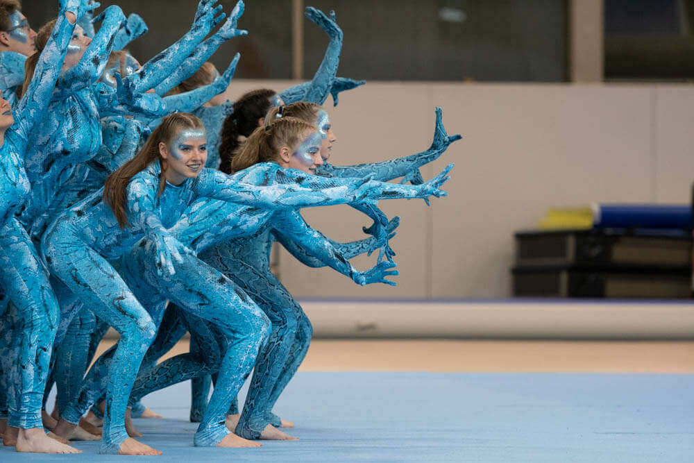 Mädchen in blauen Anzügen stehen links im Bild in Formation mit ausgestreckten Händen