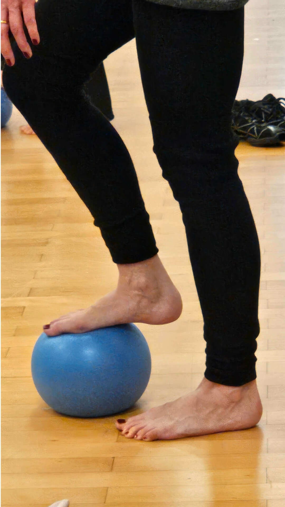 eine Person mit einer schwarten Sporthose ist barfuß stehend von der Hüfte abwärts zu sehen. Ein Bein steht am Boden, das andere Bein ist leicht gebeugt und steht mit dme Vorderfuß auf einem Gymnastikball.