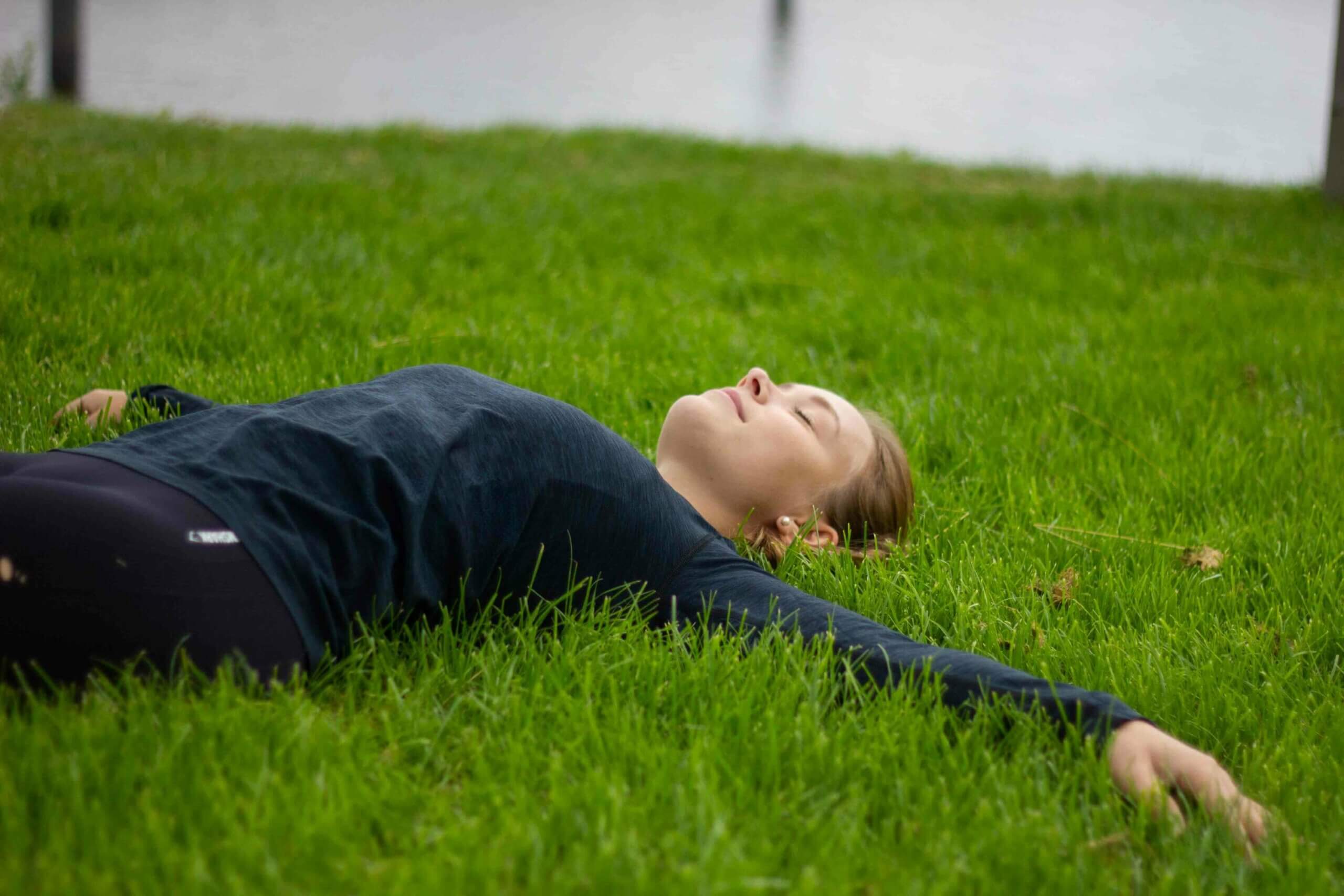 Ein Frau liegt entspannt mit ausgebreiteten Armen und geschlossenen Augen auf einer Rasenfläche
