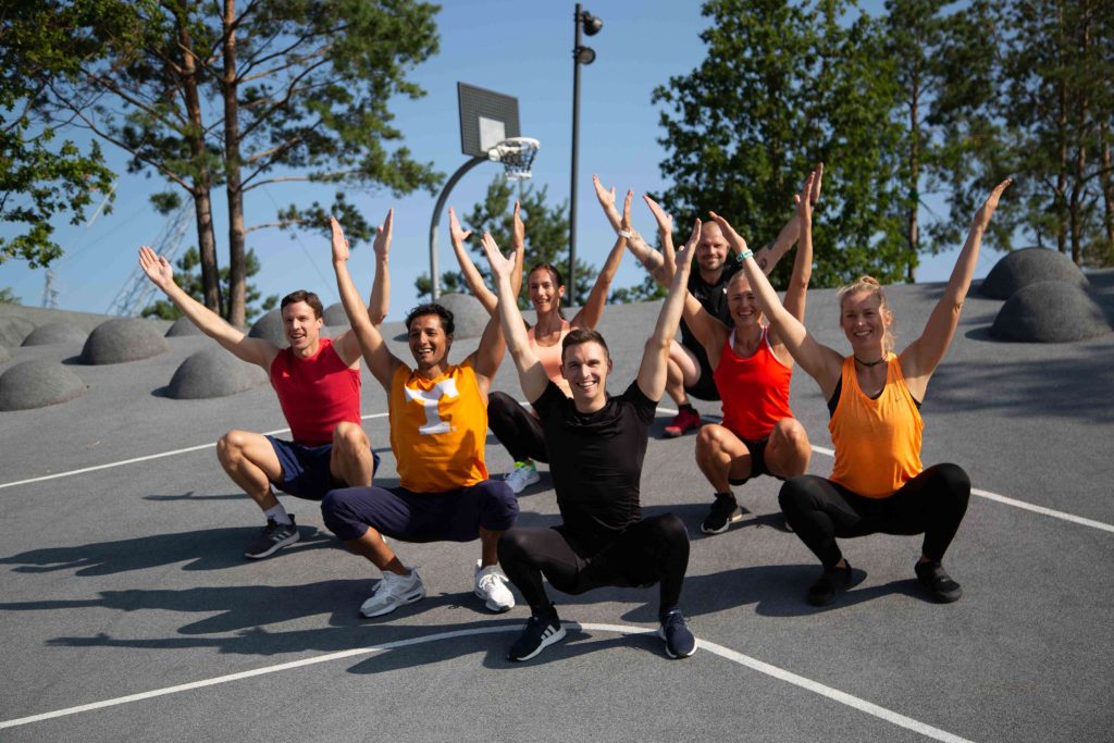 Fitness in der Gruppe macht besonders Spaß: 7 Sportler*innen sitzen in der Hocke und strecken die Arme nach oben.