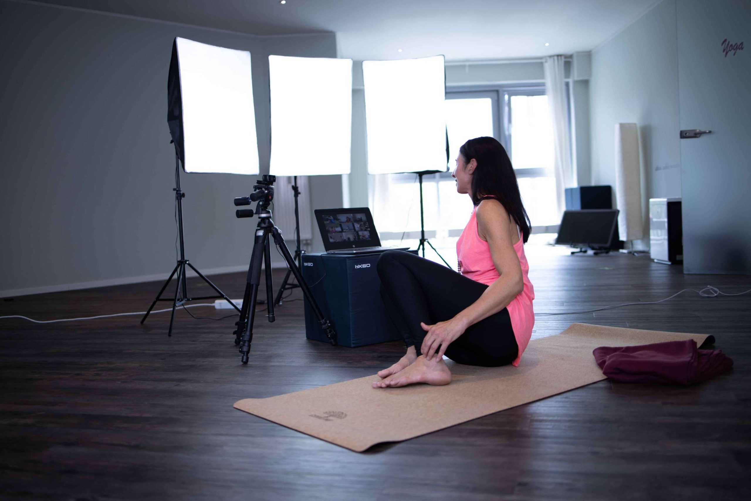 Referentin bei Online-Unterricht. Sie sitzt vor drei Lichtdisplays und einer Kamera auf einem Stativ auf ihrer Yogamatte mit Blick auf ihren ausgeklappten Laptop