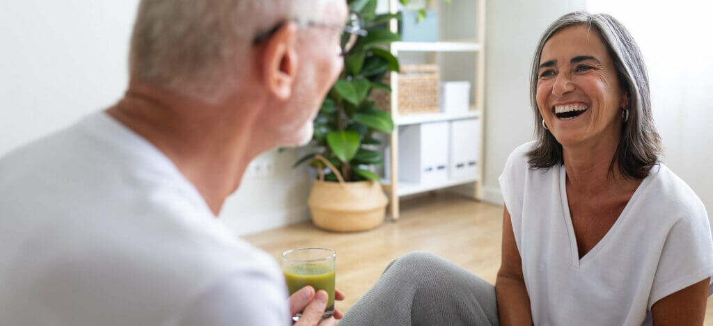 zwei ältere Menschen sitzen gemütlich zusammen und lachen, beide halten ein Getränk